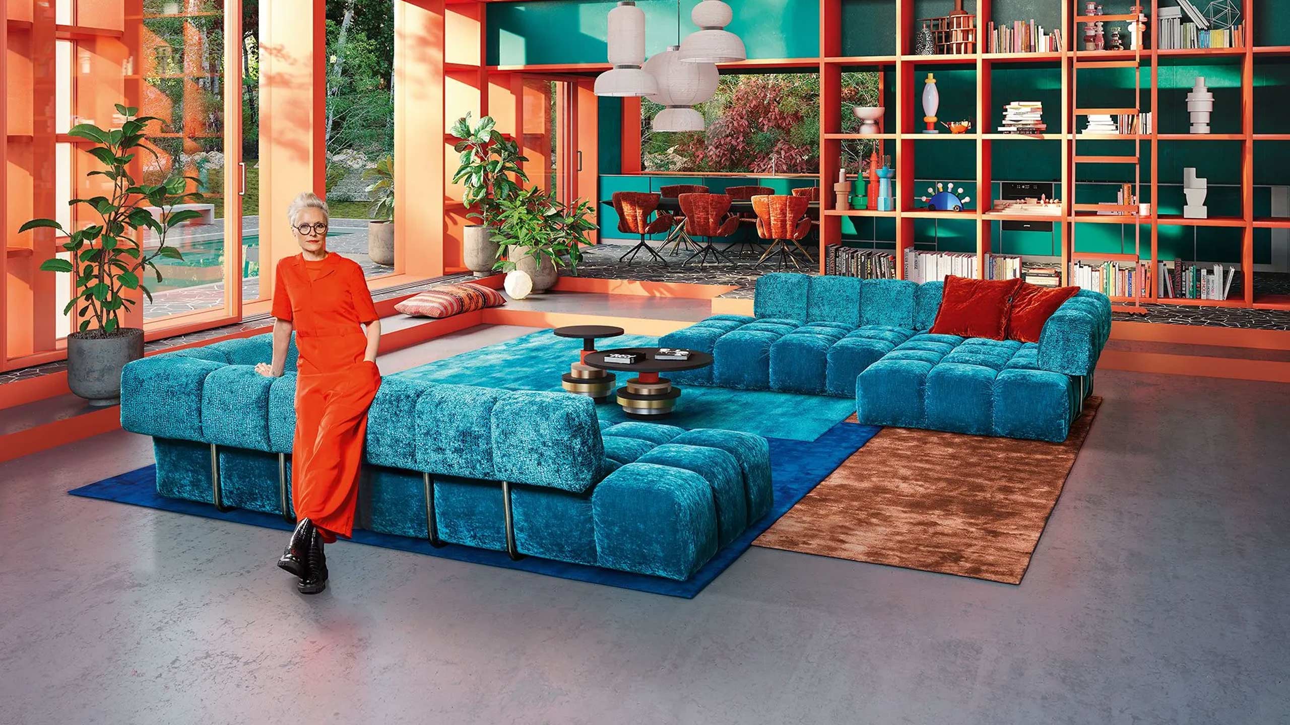 frau in orange steht gelehnt am bretz edgy big sofa in blau in einem großen Wohnzimmer mit bodentiefen fenstern